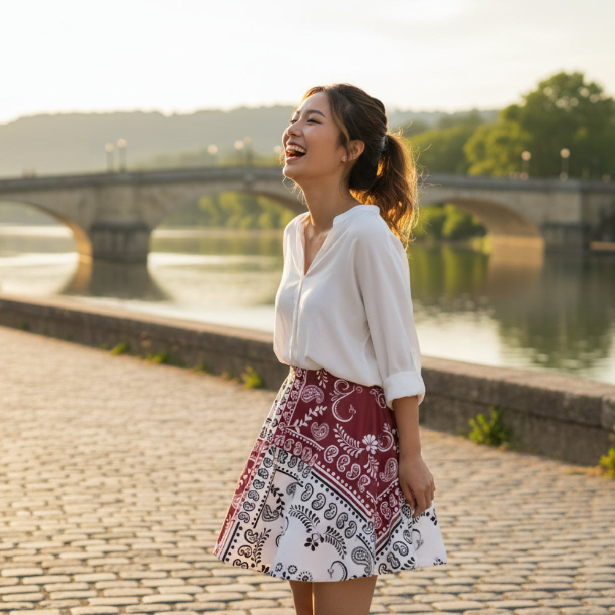 A beautiful women standing on a waterfront with a bridge in the background. She is wearing the paisley motif bandana skirt that is maroon and White, with both white and Black bandana on it. 